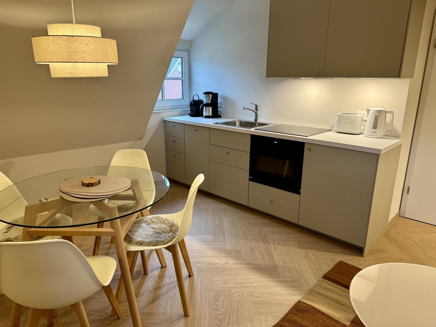 Et moderne køkken med et glasbord, hvide stole, indbygget ovn og kaffemaskine.