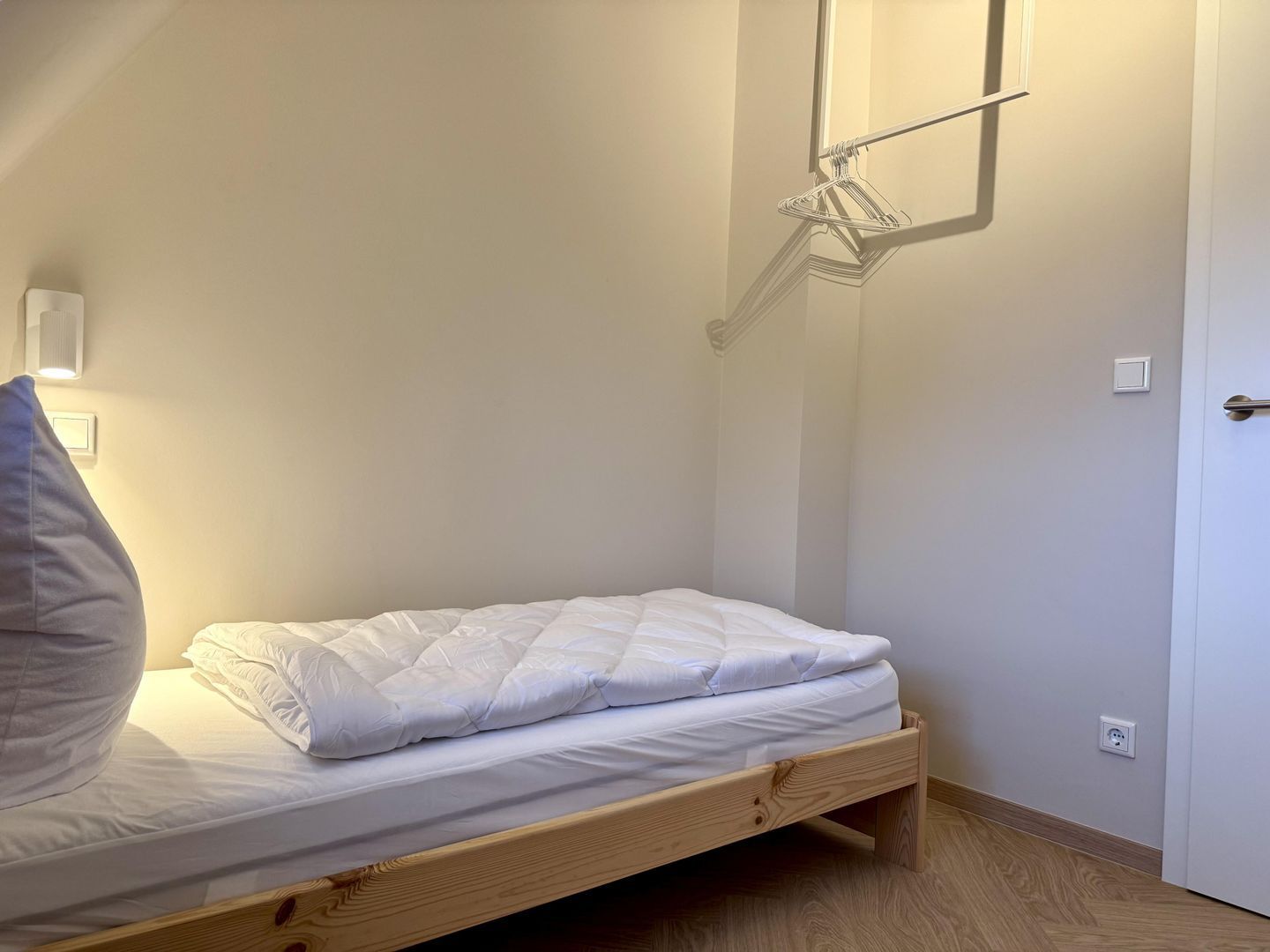 Ein Einzelbett mit Holzrahmen, einer weißen Matratze und einem grauen Kopfkissen. Eine Wandleuchte befindet sich über dem Bett und eine Tür ist rechts zu sehen.