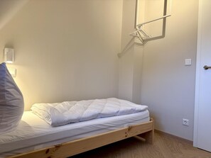 Ein Einzelbett mit Holzrahmen, einer weißen Matratze und einem grauen Kopfkissen. Eine Wandleuchte befindet sich über dem Bett und eine Tür ist rechts zu sehen.