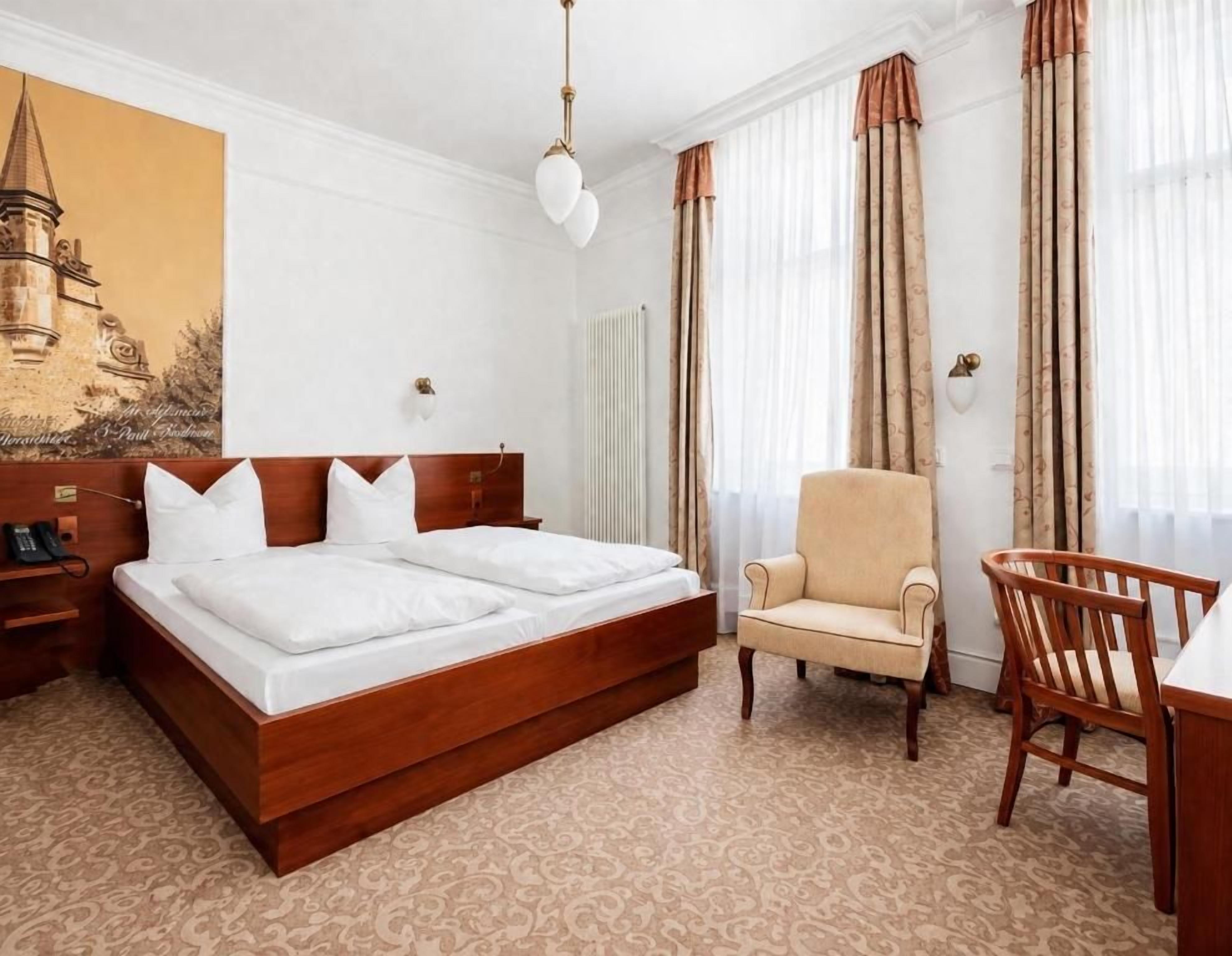 A hotel room with a double bed, a chair, a desk, and a large window with curtains.