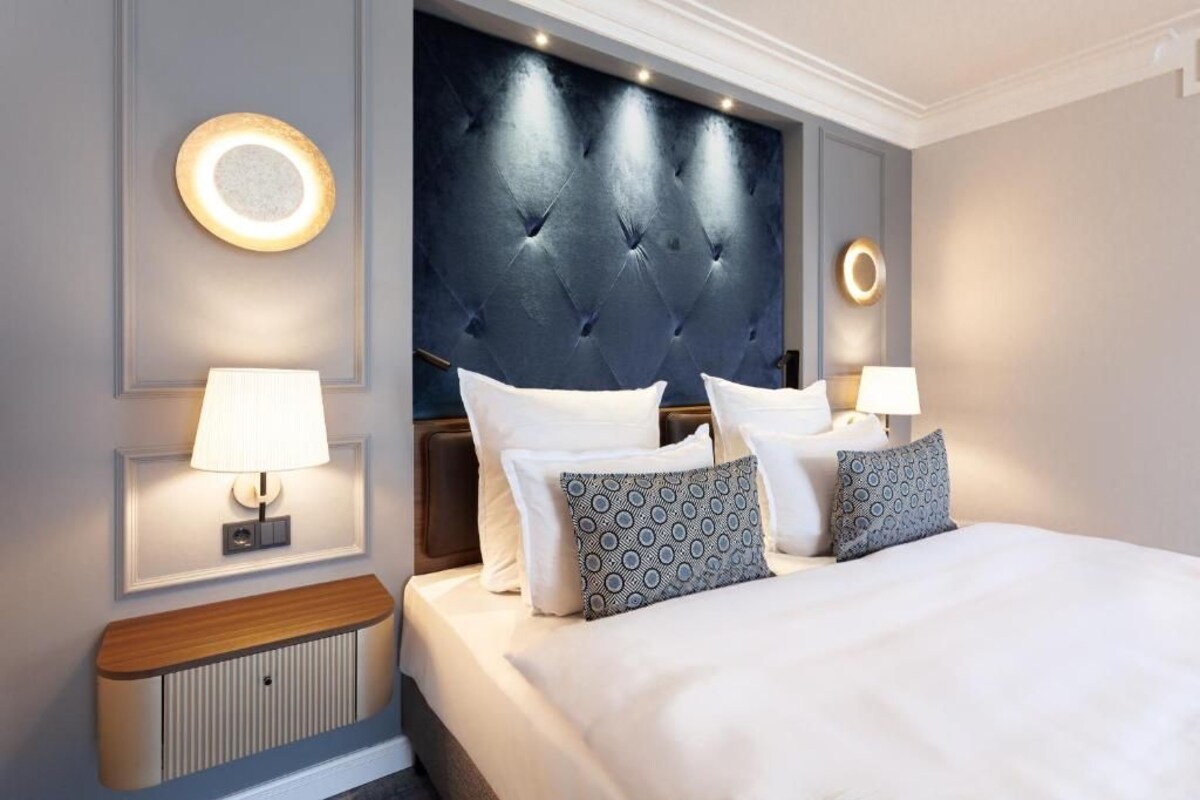 A hotel room with a tufted headboard, two bedside lamps, a wooden bedside table, and a large bed with patterned pillows.