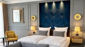 A modern hotel room with a large bed, a sofa, two bedside tables with lamps, a chair, and a large wall-mounted headboard. - Hotel Fürstenhof (Bad Pyrmont)