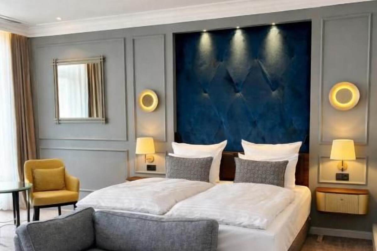 A modern hotel room with a large bed, a sofa, two bedside tables with lamps, a chair, and a large wall-mounted headboard.