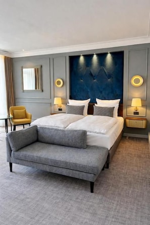 A modern hotel room with a large bed, a sofa, two bedside tables with lamps, a chair, and a large wall-mounted headboard. - Hotel Fürstenhof (Bad Pyrmont)