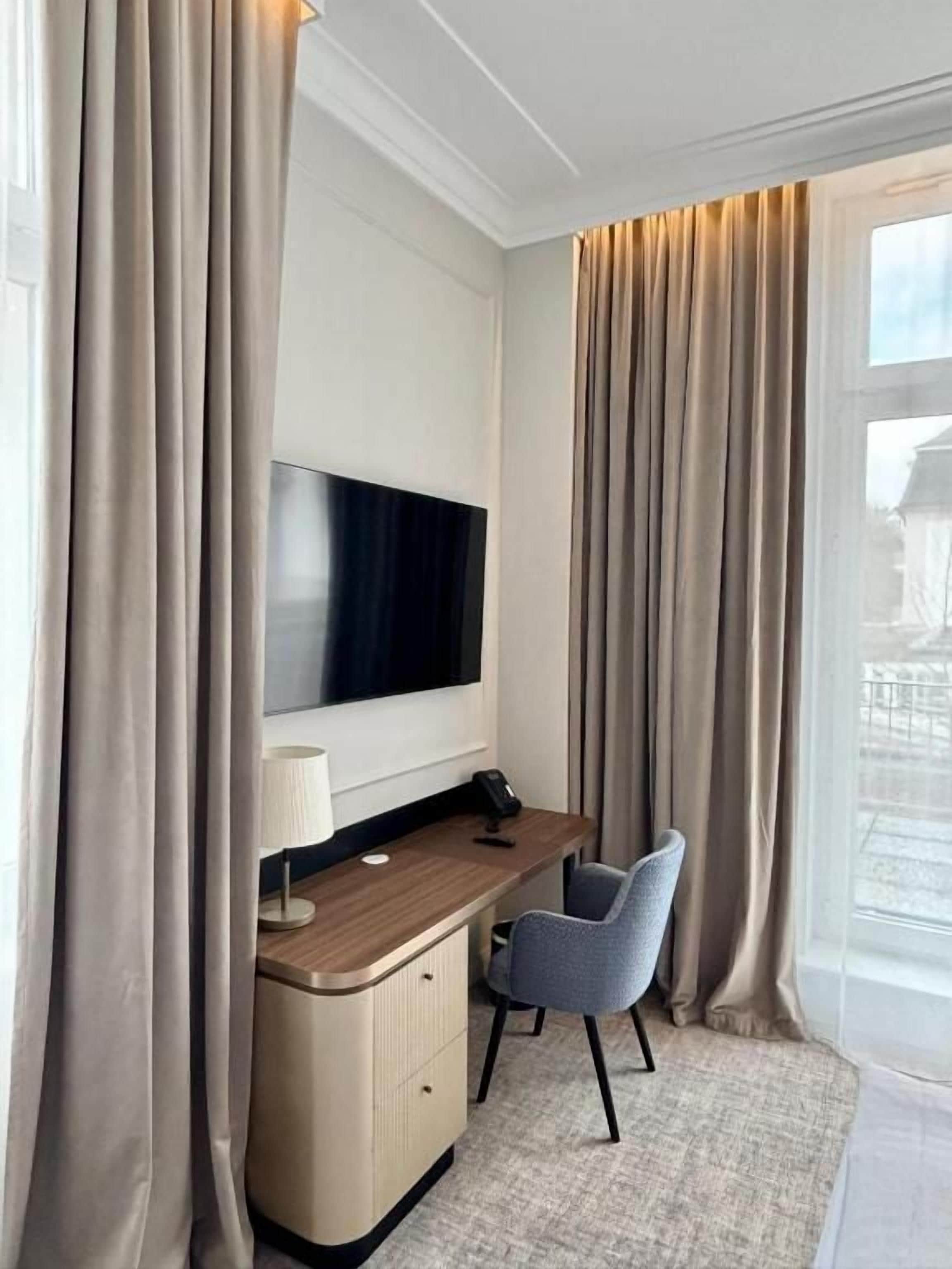 Habitación de hotel con una televisión de pantalla plana montada en la pared, un escritorio con lámpara, una silla y un ventanal con cortinas.