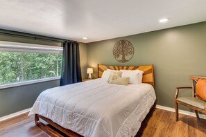 Ein Schlafzimmer mit einem großen Bett, einem Holz-Kopfteil, einem Nachttisch mit Lampe, einem Sessel und einem Fenster mit Vorhängen.