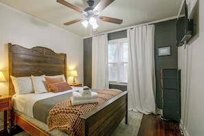 A neatly arranged bedroom with a wooden headboard, a bed with white and patterned bedding, a bedside lamp, a window with curtains, and a ceiling fan. - East Memphis Duplex Near Midtown & Downtown (Memphis)