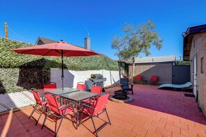 Un patio exterior con suelo de ladrillo, sillas rojas, una mesa, una parrilla y un paraguas rojo.