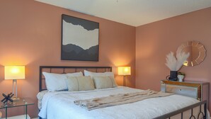 A neatly made bed with a white duvet and multiple pillows, a bedside table with a lamp, a wall-mounted abstract painting, and a round mirror with a decorative frame. - Hot Tub, Water View! Largo Home 10 Mi to Beaches (Largo)