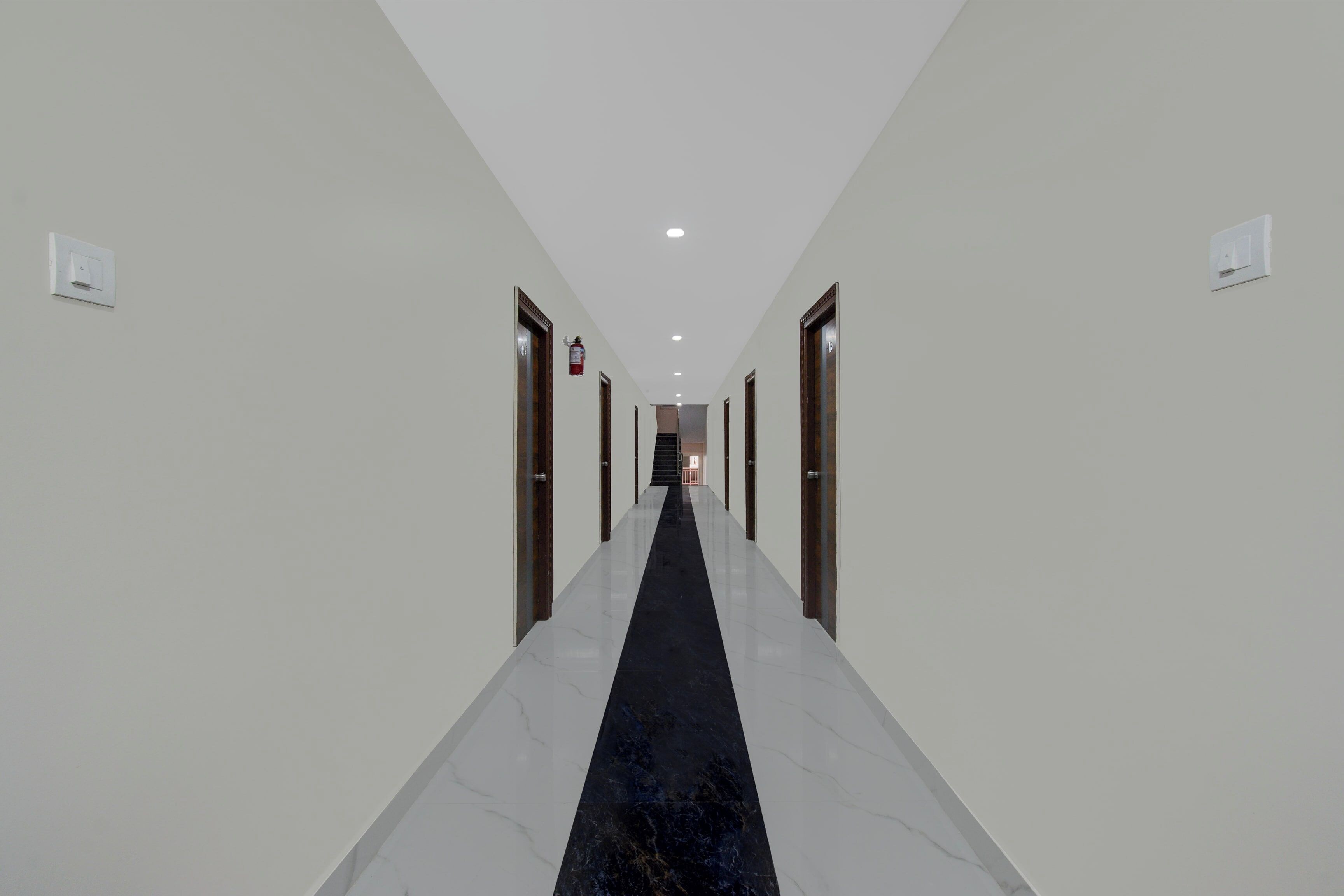 Un couloir d’hôtel avec un tapis noir, des murs blancs et des portes de chaque côté.