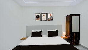 Quarto de hotel com uma cama grande, duas almofadas pretas, mesa de cabeceira com abajur, guarda-roupa e três obras de arte emolduradas na parede.