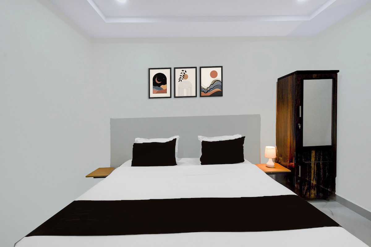 A hotel room with a large bed, two black pillows, a nightstand with a lamp, a wardrobe, and three framed artworks on the wall.