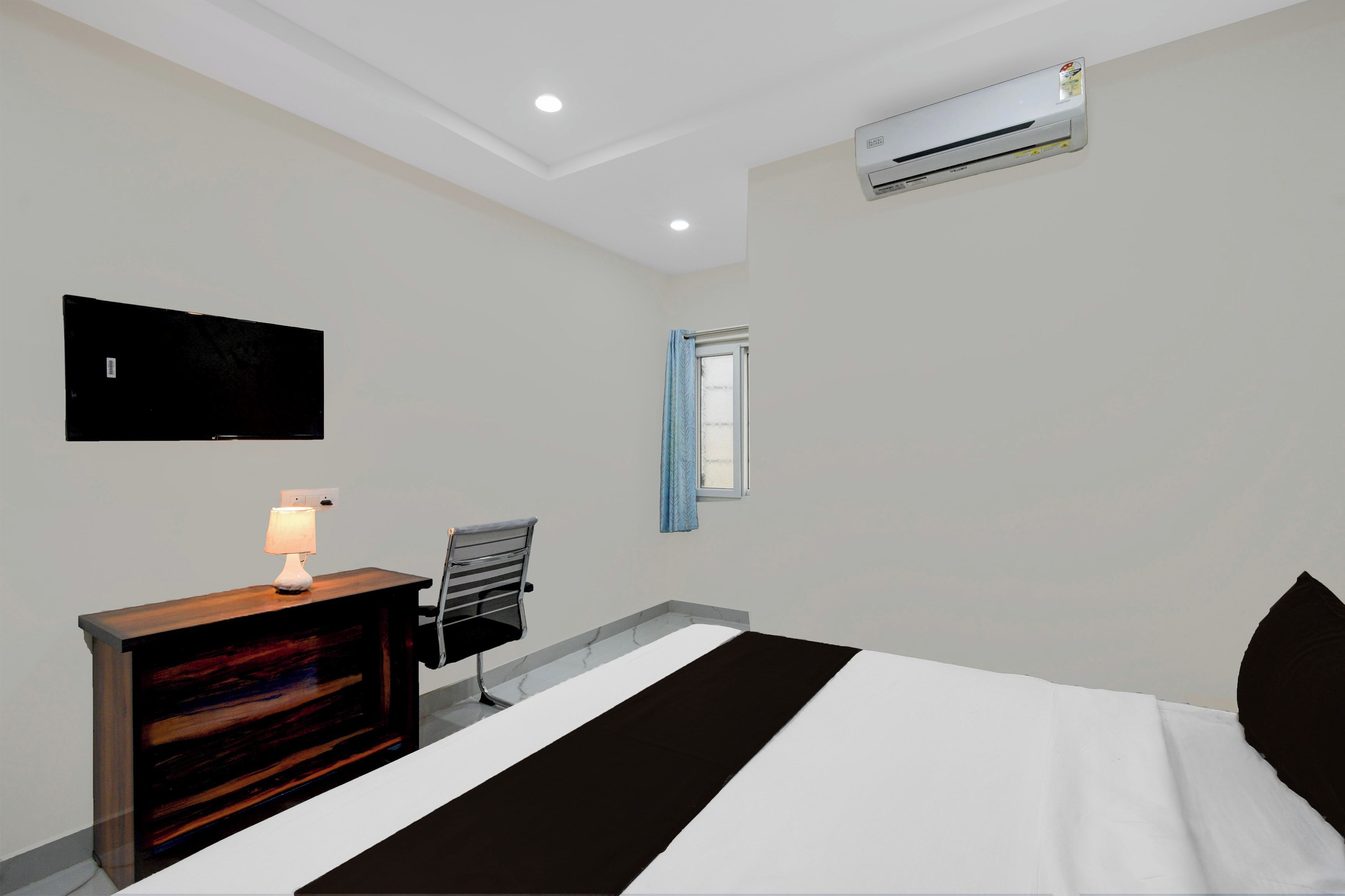Una camera d'albergo con un letto, una scrivania con una lampada, una sedia, una televisione a muro e un condizionatore d'aria.