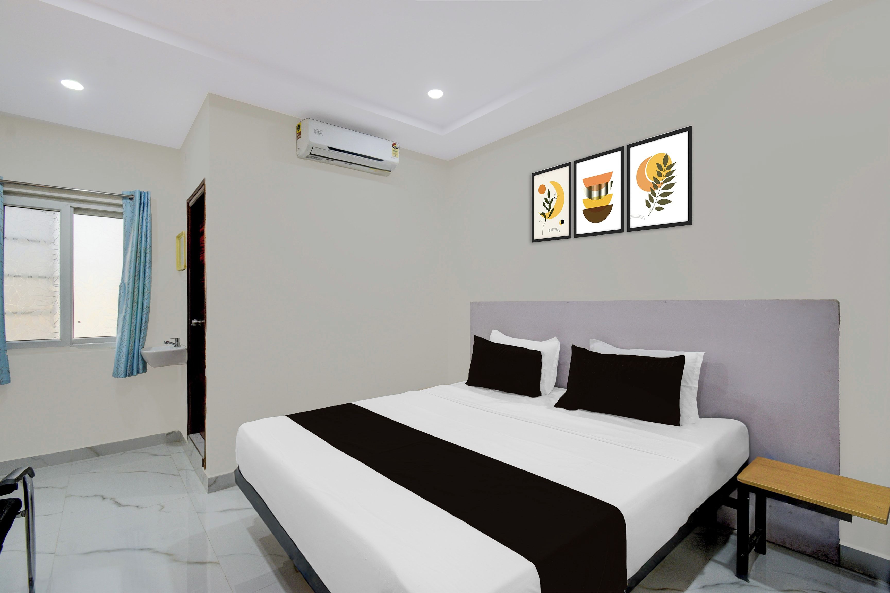 A hotel room with a bed, a bedside table, a window with curtains, an air conditioning unit, and three framed artworks on the wall.