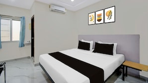 Quarto de hotel com cama, mesa de cabeceira, janela com cortinas, aparelho de ar condicionado e três obras de arte emolduradas na parede.