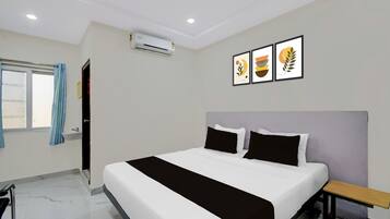 A hotel room with a bed, a bedside table, a window with curtains, an air conditioning unit, and three framed artworks on the wall.