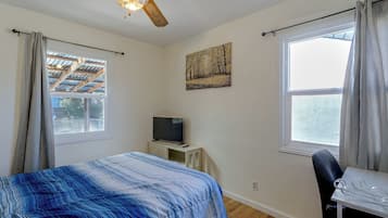 Un dormitorio con una cama, un televisor sobre una mesita, un ventilador de techo y una ventana con cortinas.