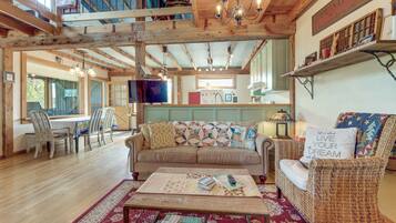 A cozy interior with a wooden ceiling, a dining area with chairs and a table, a living room with a sofa and a coffee table, and a kitchen in the background.