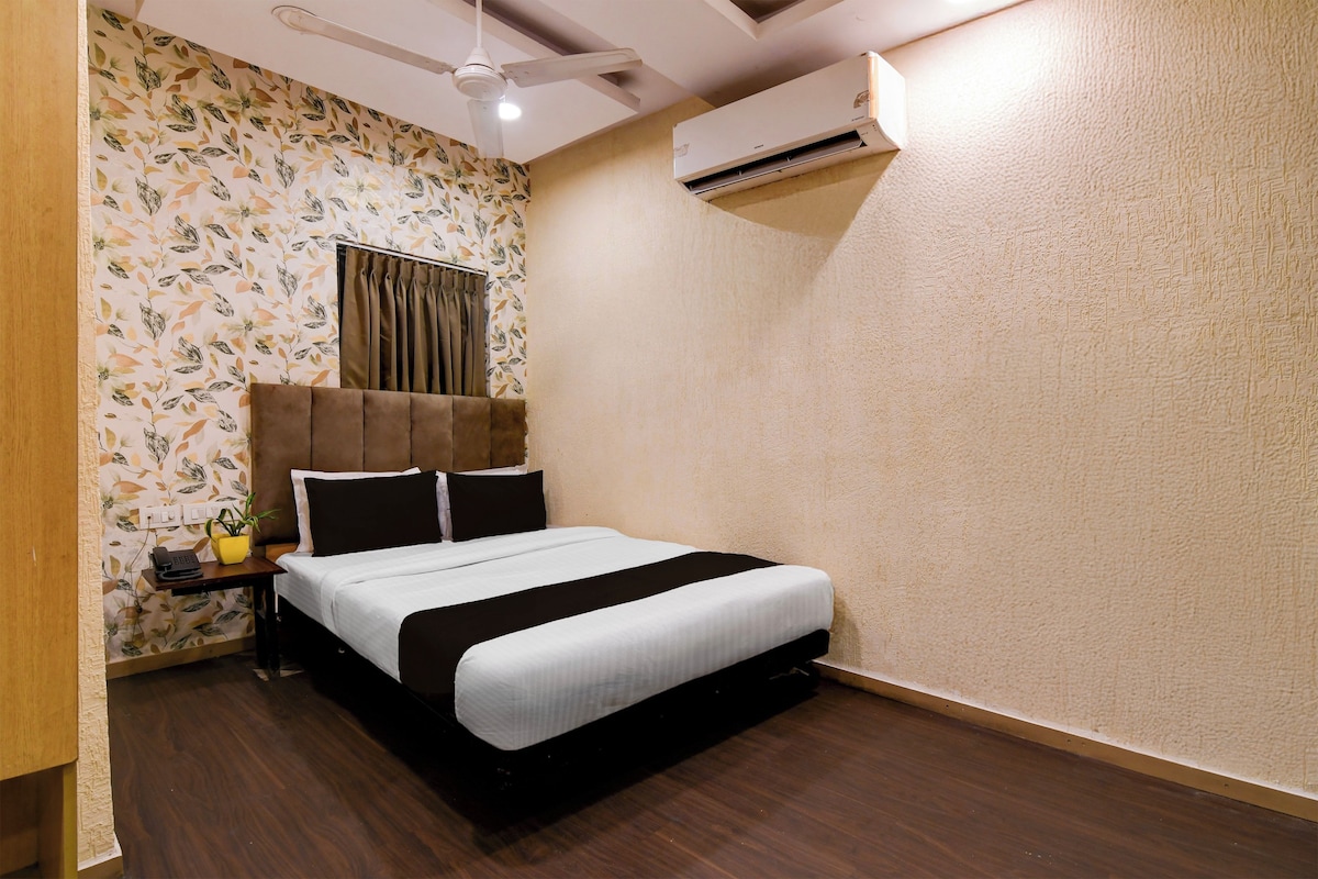 A hotel room with a bed, a bedside table with a phone, a plant, and a window with curtains.