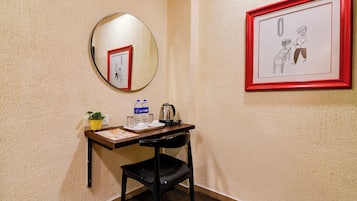 Quarto nos cantos com escrivaninha, cadeira, quadro emoldurado, espelho redondo e planta em vaso.