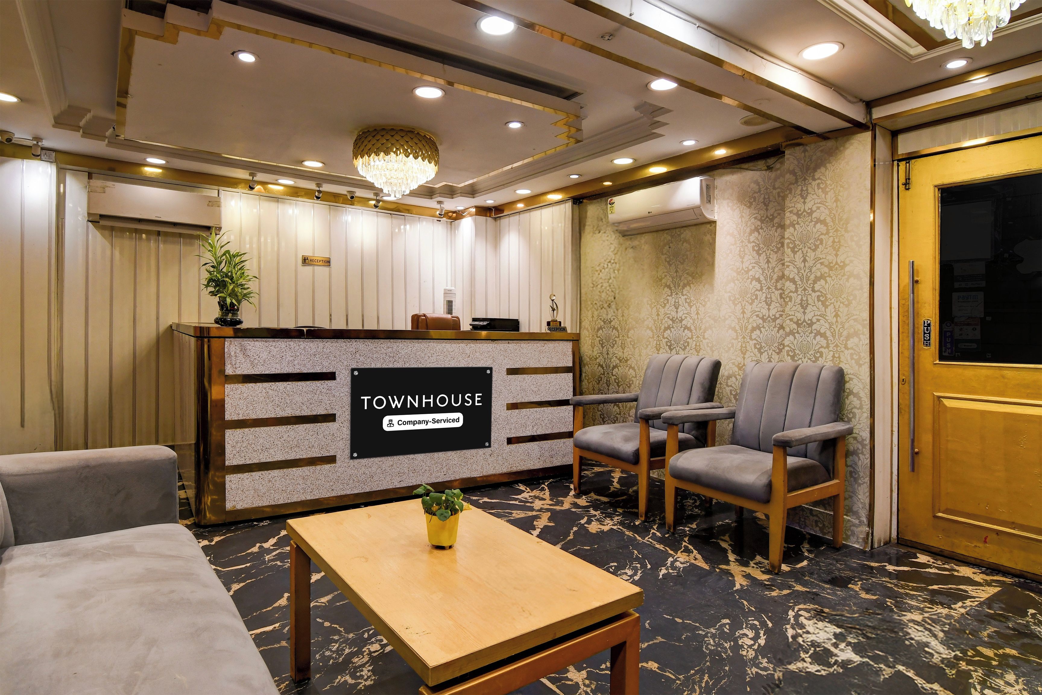 Un área de recepción con un letrero que dice 'TOWNHOUSE Company-Serviced', dos sillones grises, una mesa de madera con una planta y una araña.
