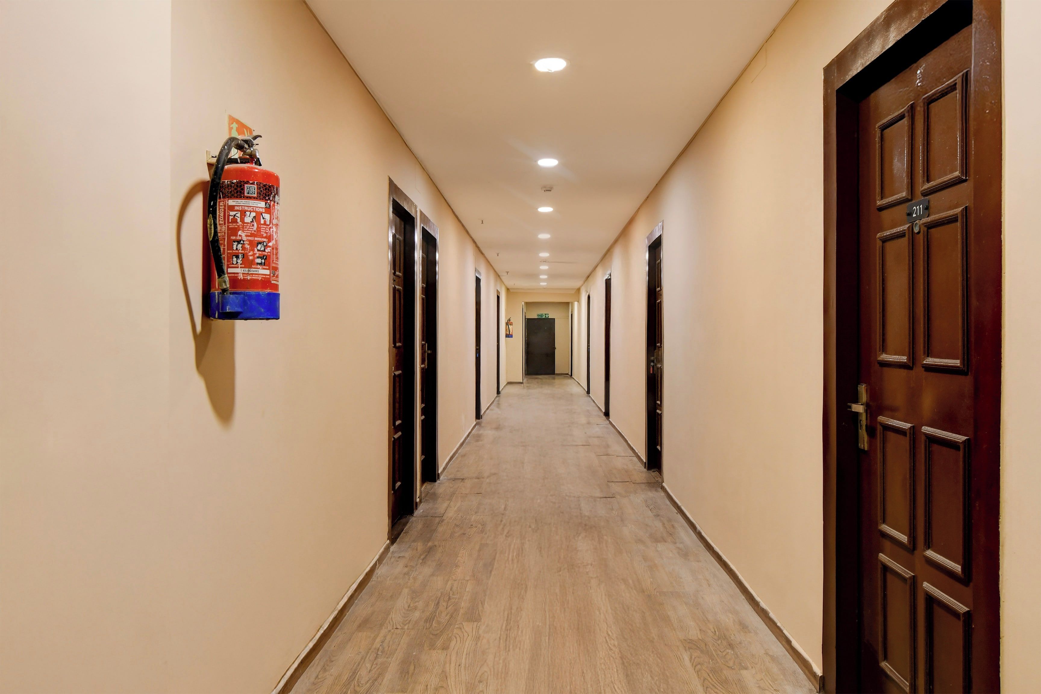 A hotel corridor with wooden flooring, doors on both sides, and a fire extinguisher mounted on the wall.