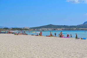 A beach with people sunbathing and swimming, a pier in the background, and a hilly landscape.
