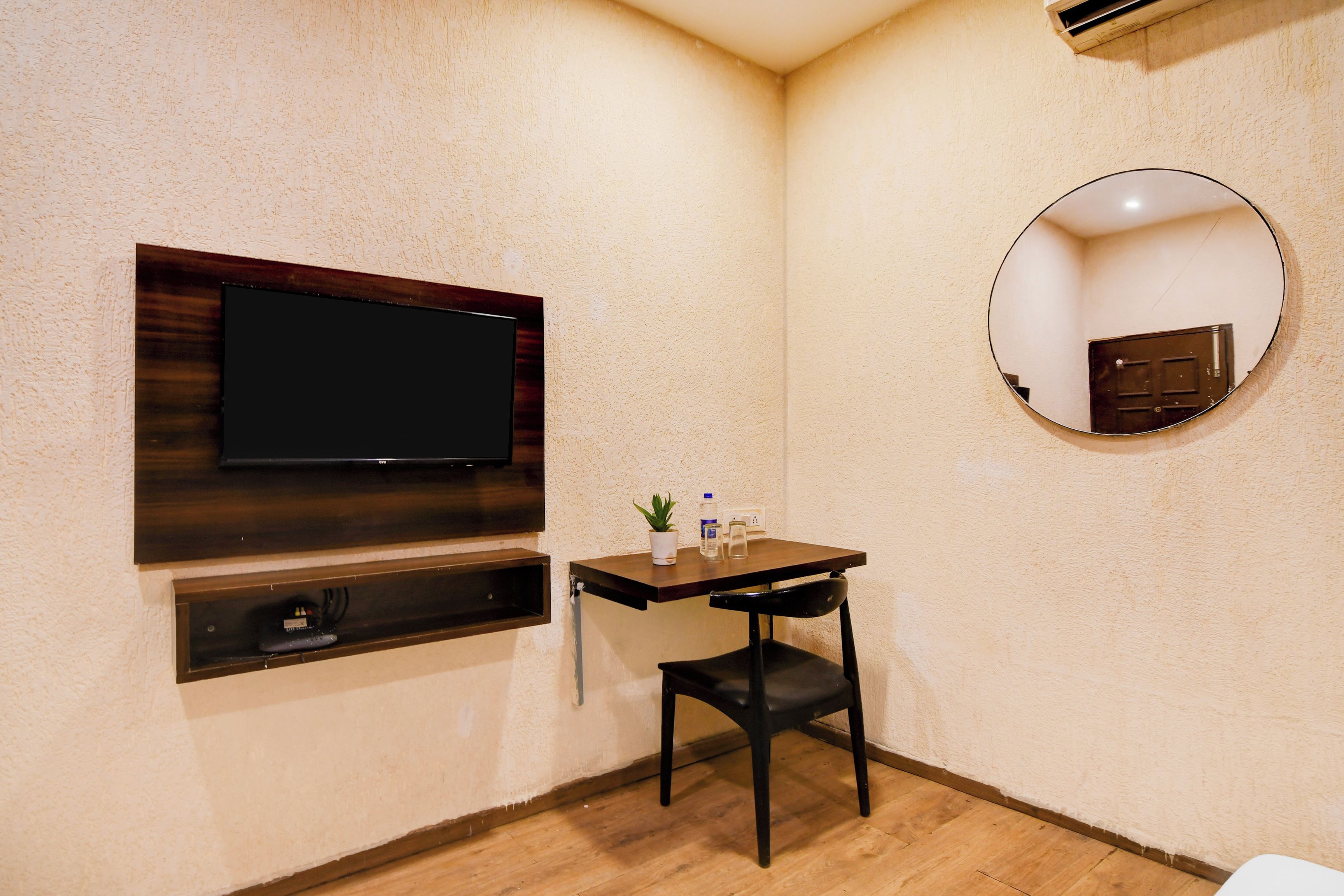 A small room with a flat-screen TV mounted on the wall, a small table with a chair, a potted plant, and a round mirror.