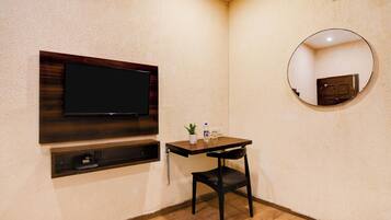 A small room with a flat-screen TV mounted on the wall, a small table with a chair, a potted plant, and a round mirror.