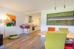 A modern kitchen with white cabinets, a dining area with a wooden table and colorful chairs, and a painting on the wall. - Alpenflair holiday apartment 227 (Oberstdorf)