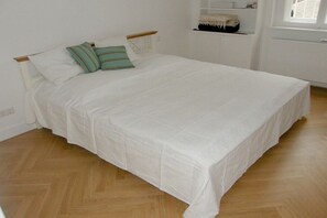 A single-bed room with a wooden floor, white bedding, and a small shelf with items. - Villa Hoyerberg Lake (Lindau)