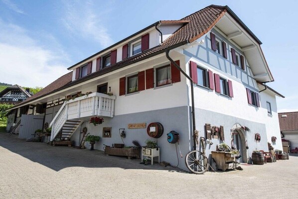 Ein zweistöckiges Gebäude mit einer Treppe, die zu einem Balkon führt, umgeben von Topfpflanzen und Gartenmöbeln.