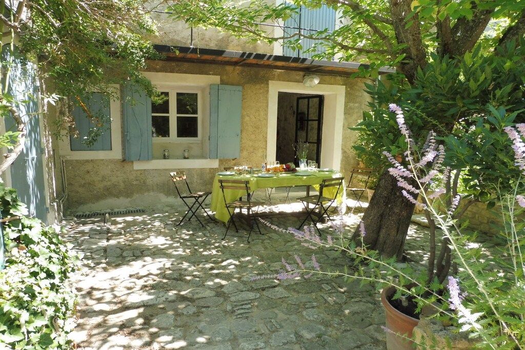 A stone-paved courtyard with a table set for dining, surrounded by greenery and a building with blue shutters.