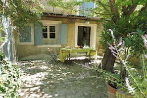 A stone-paved courtyard with a table set for dining, surrounded by greenery and a building with blue shutters.
