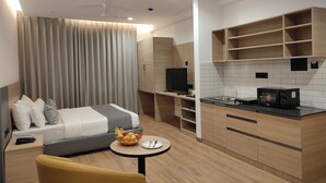 Moderni hotellihuone, jossa on sänky, pieni pyöreä pöytä hedelmäkulhon kanssa, sohva ja pieni keittiö, jossa on mikroaaltouuni.