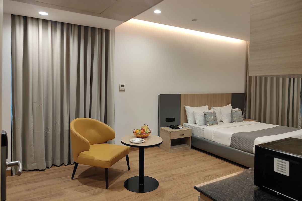 Une chambre d’hôtel comprenant un lit, une table de chevet, une chaise, une petite table ronde avec un panier de fruits et un panneau de commande au mur.