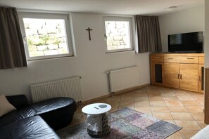 Ein Wohnzimmer mit einem schwarzen Ledersofa, einem kleinen runden Tisch mit weißer Tischplatte, einem Fernseher auf einer Holzkommode und zwei mit Vorhängen versehenen Fenstern.