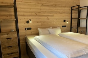 Quarto com parede de madeira, cama com roupa de cama branca, duas mesas de cabeceira e luminárias de leitura fixadas na parede.