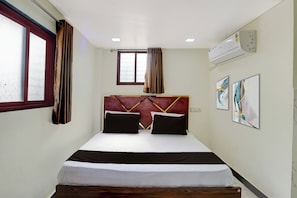 Une chambre d’hôtel avec un lit, une fenêtre avec des rideaux, un climatiseur fixé au mur et deux œuvres d’art encadrées.