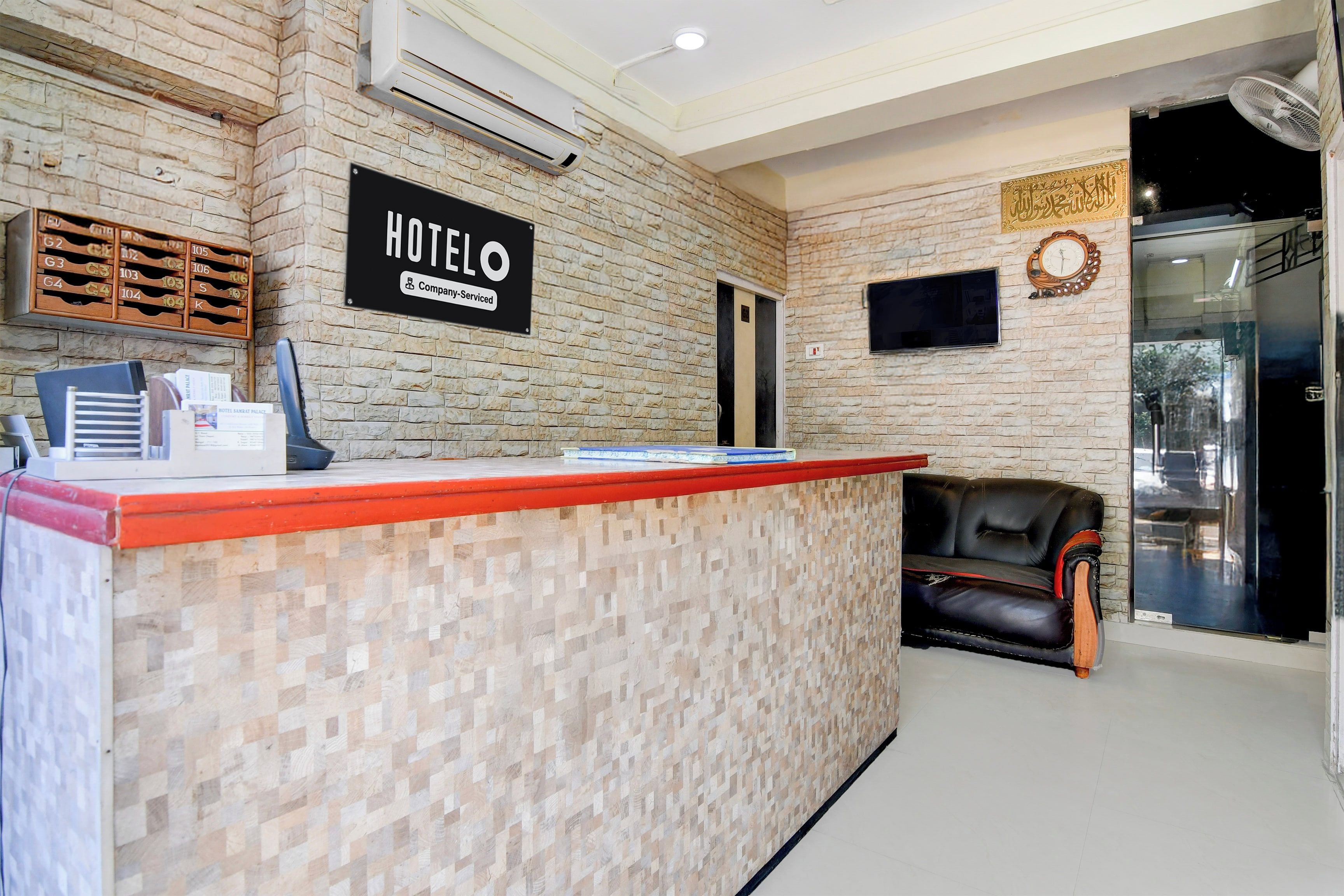 Ein Hotelrezeptionstresen mit einem Schild, das anzeigt, dass es sich um ein betriebliches Hotel handelt. Es gibt einen Empfangsbereich mit einer Ledersofa und einem an der Wand montierten Fernseher.
