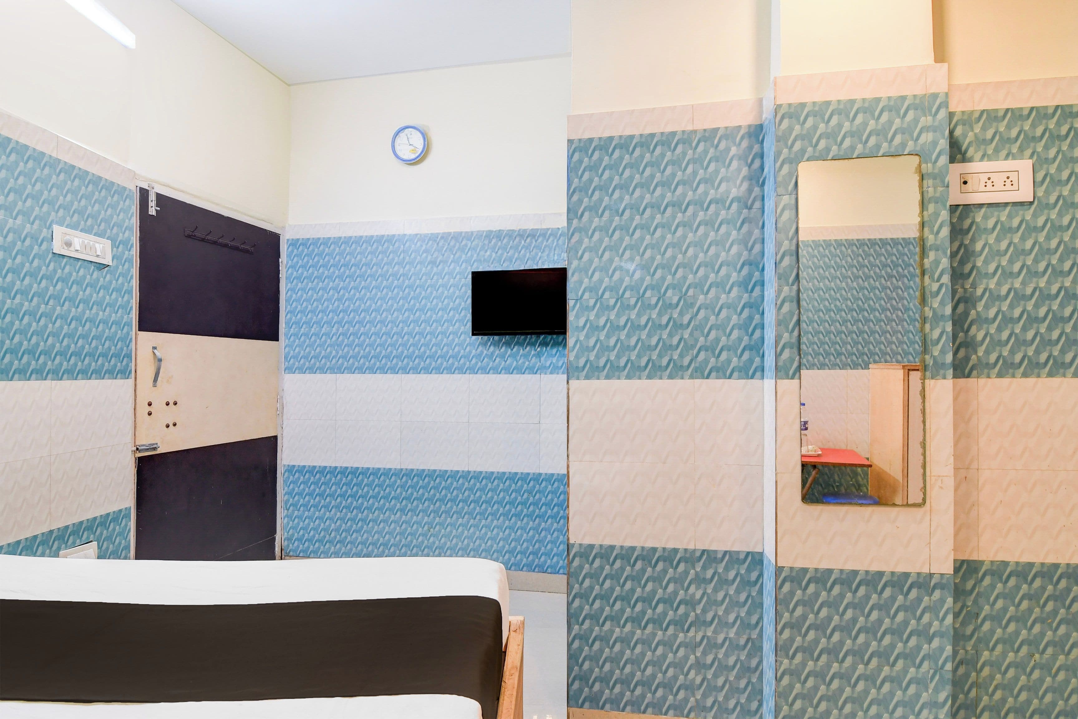 A hotel room with a bed, a wall-mounted TV, a clock, and a power outlet.