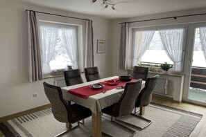 Uma sala de jantar com uma mesa posta para seis pessoas, rodeada por cadeiras, e janelas em ambos os lados.