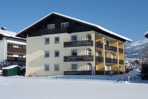 Hotel de vários andares com varandas, telhado coberto de neve e montanhas ao fundo.
