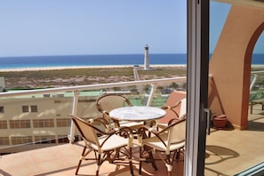 A balcony with a table and chairs, overlooking a beach and a lighthouse. - Great views || Morro Jable, Fuerteventura (Morro Jable, Fuerteventura)