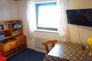A room with a window, a television mounted on the wall, a wooden bookshelf, a table with a colorful tablecloth, and a red chair. - Holiday home in Zandt with swimming pool (Zandt)