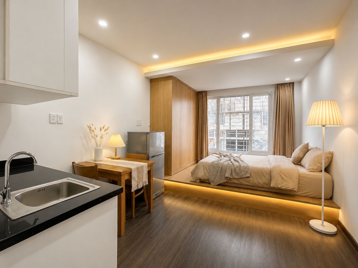 A modern hotel room with a kitchenette, a bed with a view, and a bedside lamp.