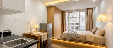 A modern hotel room with a kitchenette, a bed with a view, and a bedside lamp.