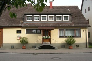Ein zweistöckiges Gebäude mit einem braunen Ziegeldach, einer hölzernen Haustür und einer kleinen Veranda mit Stufen. Vor dem Eingang stehen je ein Blumentopf auf jeder Seite.
