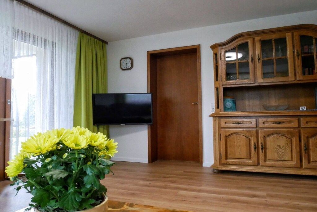 A living room with a flat-screen TV mounted on the wall, a wooden cabinet with glass doors, and a potted plant with yellow flowers.