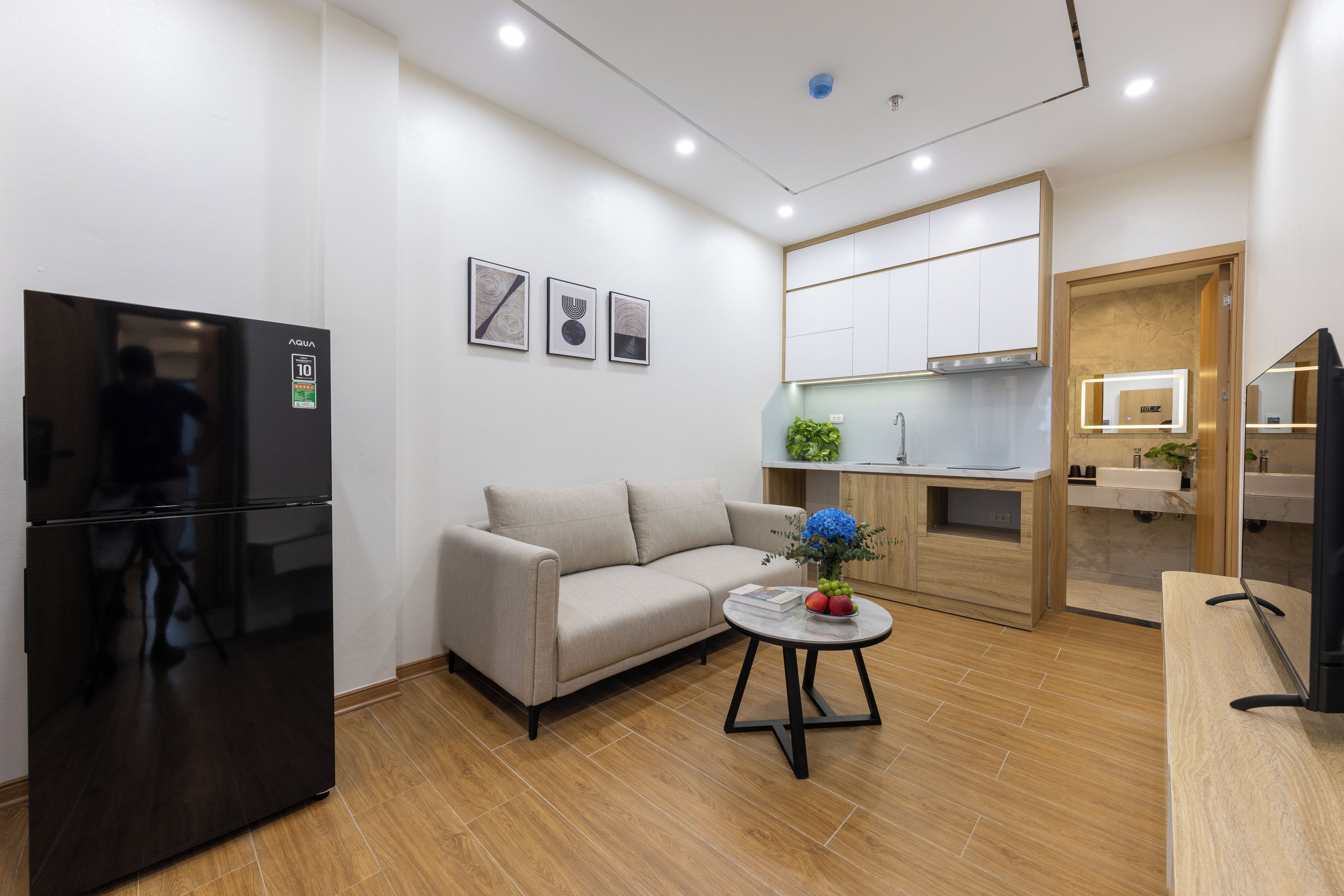 Una sala moderna con un sofá, una mesa de centro con un florero, un refrigerador negro y una cocina con armarios y un lavamanos.